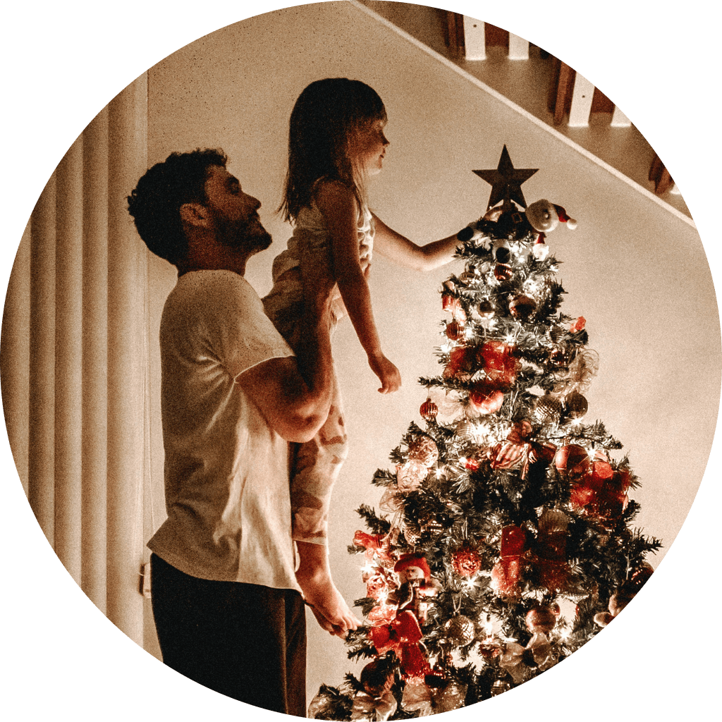 Mies nostaa pikkutyttöä laittamaan tähden joulukuusen latvaan. Kuusi on koristeltu punaisin ja kultaisin koristein, ja se loistaa lämpimässä valaistuksessa. Taustalla on valkoinen seinä ja hämärästi valaistu huone.