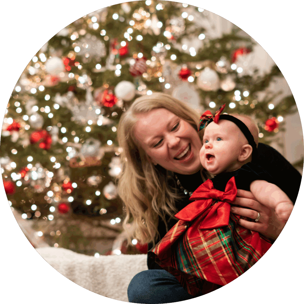 Nainen hymyilee ja pitää vauvaa sylissään joulukuusen edessä. Vauva on pukeutunut punaiseen ja vihreään mekkoon, jossa on iso rusetti.
