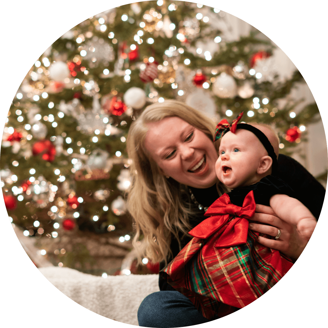 Nainen hymyilee ja pitää vauvaa sylissään joulukuusen edessä. Vauva on pukeutunut punaiseen ja vihreään mekkoon, jossa on iso rusetti.