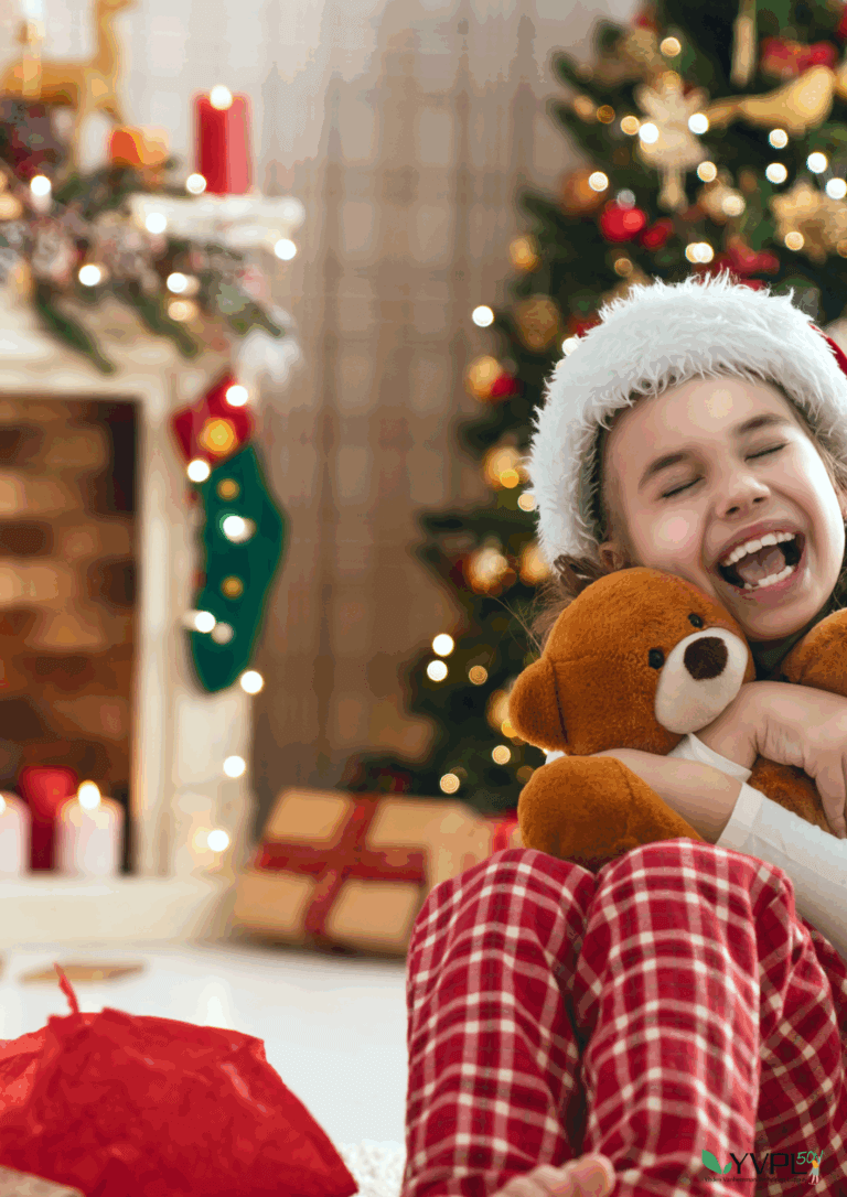 Lapsi iloisena halaten pehmolelua joulukuusen edessä, yllään tonttulakki ja ruudulliset pyjamahousut. Takana näkyy koristeellinen takka ja joululahjoja.