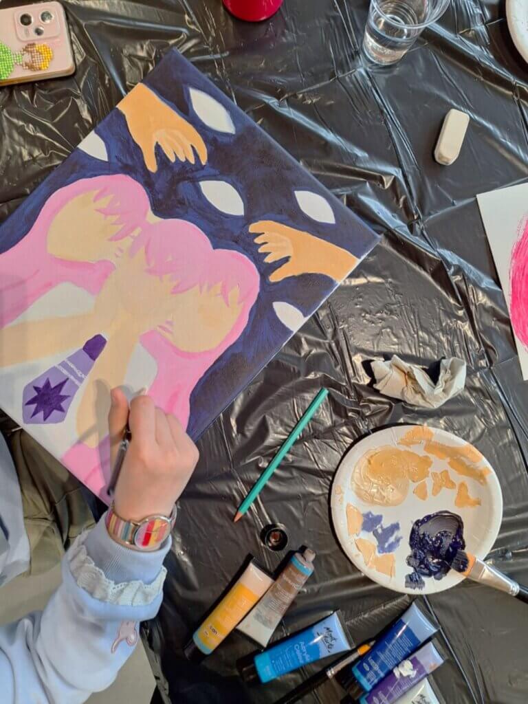 Henkilö maalaa taulua, jossa on vaaleanpunainen ja sininen kuvio kädellä. Pöydällä on kivikuvioinen matkapuhelin, pensseleitä, maalituubeja, vesilasi ja paletti, jossa on maalivärejä. Tausta on musta mu