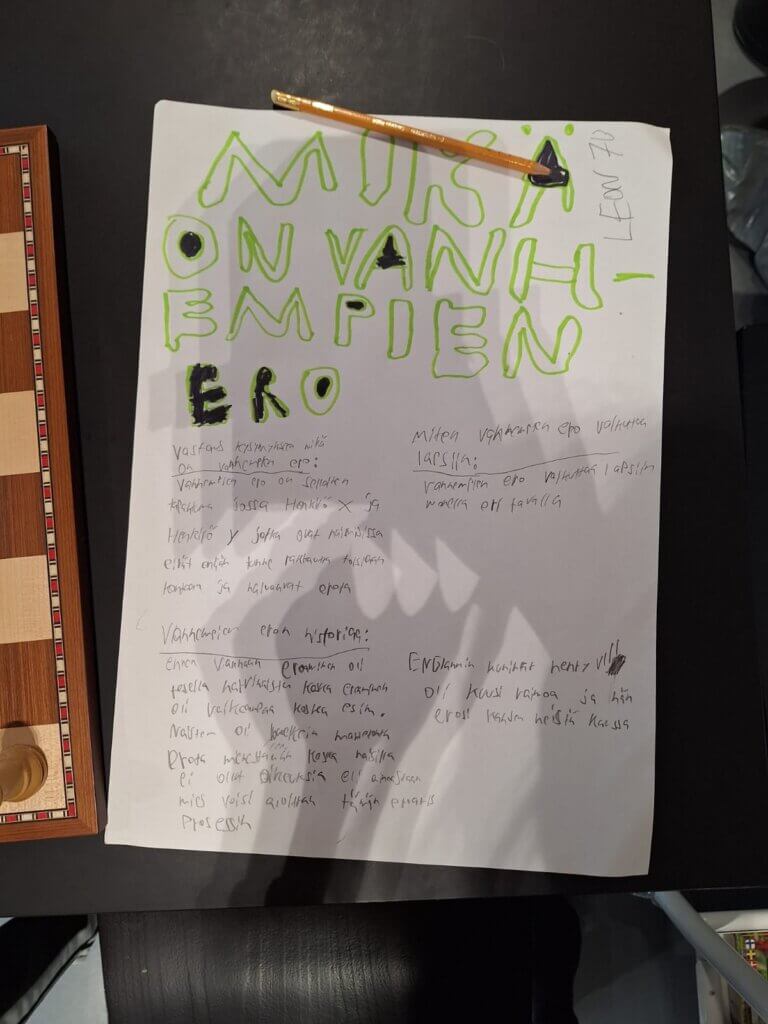 Kuvassa on paperi, jossa on käsin kirjoitettua ja suurella tekstillä otsikko MIKÄ ON VANHEMPIEN ERO. Paperilla on tekstiä sekä vihreällä että mustalla. Paperin päällä on puinen lyijykynä. Taustalla näkyy osittain auko