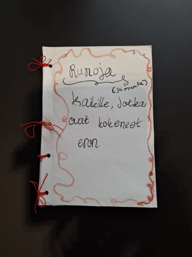 Kuvassa on käsin tehty kirja, jossa on valkoinen kansi ja punaisella nauhalla sidotut reunat. Kannessa lukee käsin kirjoitettuna: Runoja (ja muuta) kaikille, jotka ovat kokeneet eron. Leipäteksti on mustalla ja osa kehyst