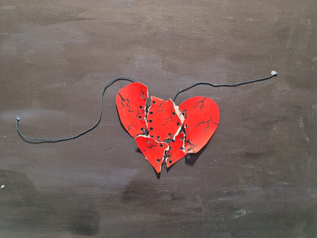 Kuvassa on punainen sydän, joka on rikkinäinen ja halkeillut. Sydän on sidottu kokoon mustalla narulla, joka kulkee kummaltakin puolelta. Tausta on yksivärinen ja neutraali.