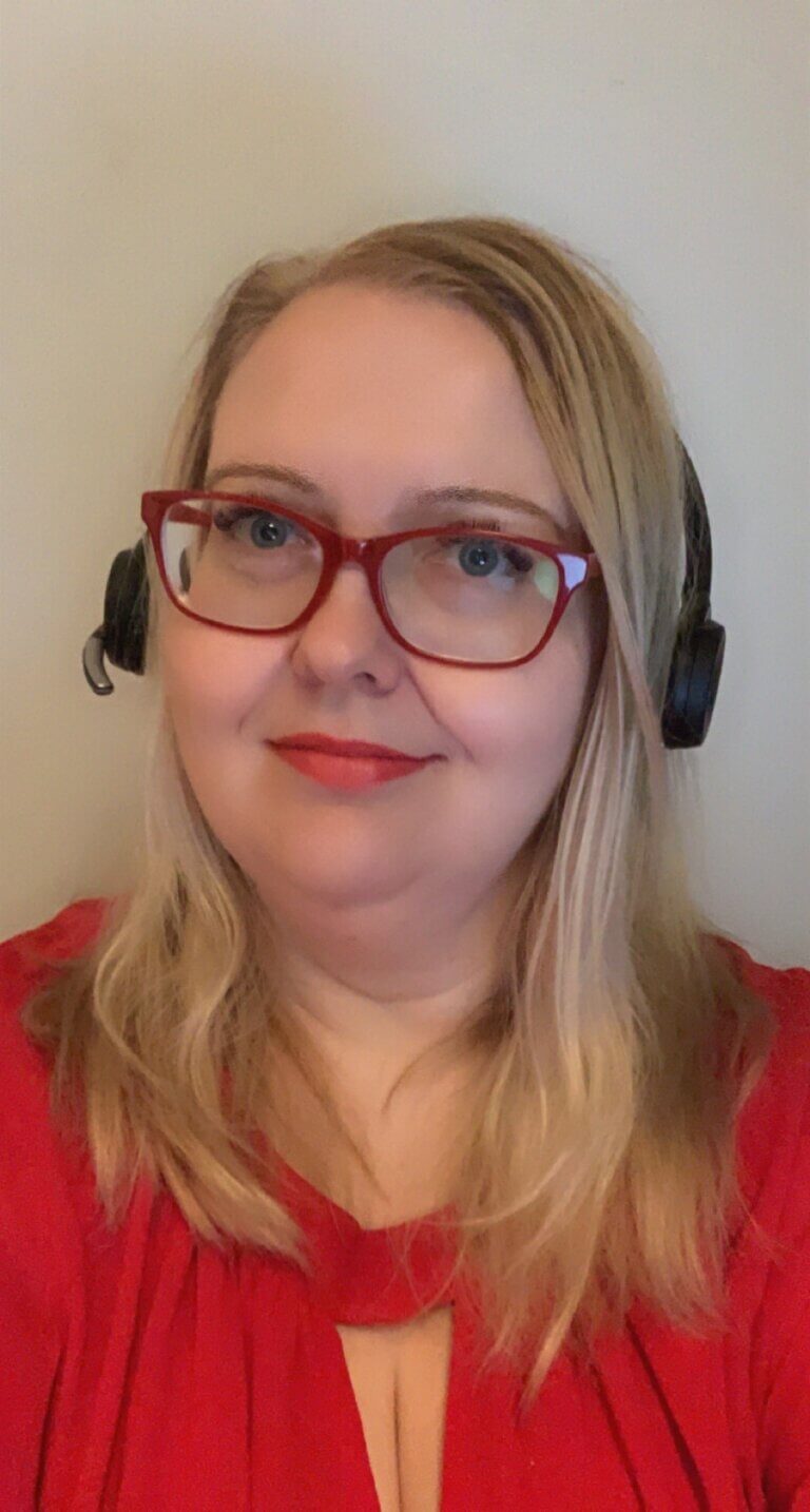 Nainen, jolla on punaiset silmälasit ja päällään punainen pusero, käyttää kuulokemikrofonia. Hän katsoo suoraan kameraan ja hymyilee hennosti.