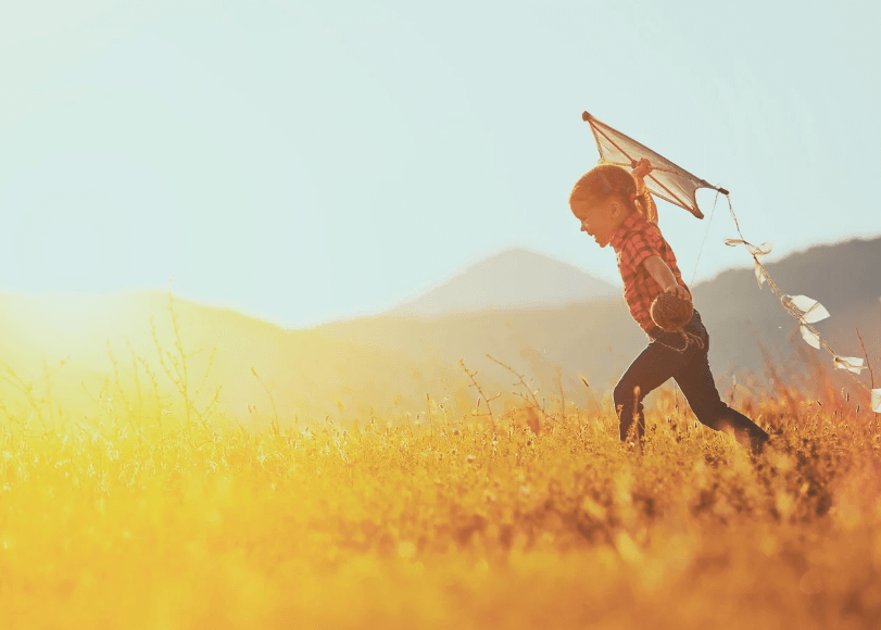 Lapsi juoksee niityllä auringonlaskussa pitäen kädessään leijaa. Taustalla näkyy vuoria ja kultainen valaistus peittää maiseman.