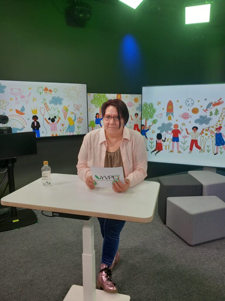 Henkilö seisoo pöydän ääressä värikkäässä studiossa, jossa on lasten piirtämiä kuvia taustalla. Hän pitelee käsissään lappua, jossa lukee YYPLSCON. Pöydällä on vesipullo.