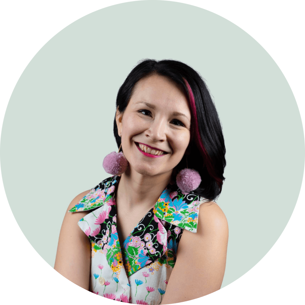 <![CDATA[Kuvassa on hymyilevä henkilö, jolla on tumma ja vaaleanpunaisilla raidoilla korostettu hiukset. Henkilöllä on suuret, vaaleanpunaiset tupsukorvakorut ja värikäs, kukkakuvioinen paita. Taustalla on vaalea ympyrämuotoinen alue.]]>