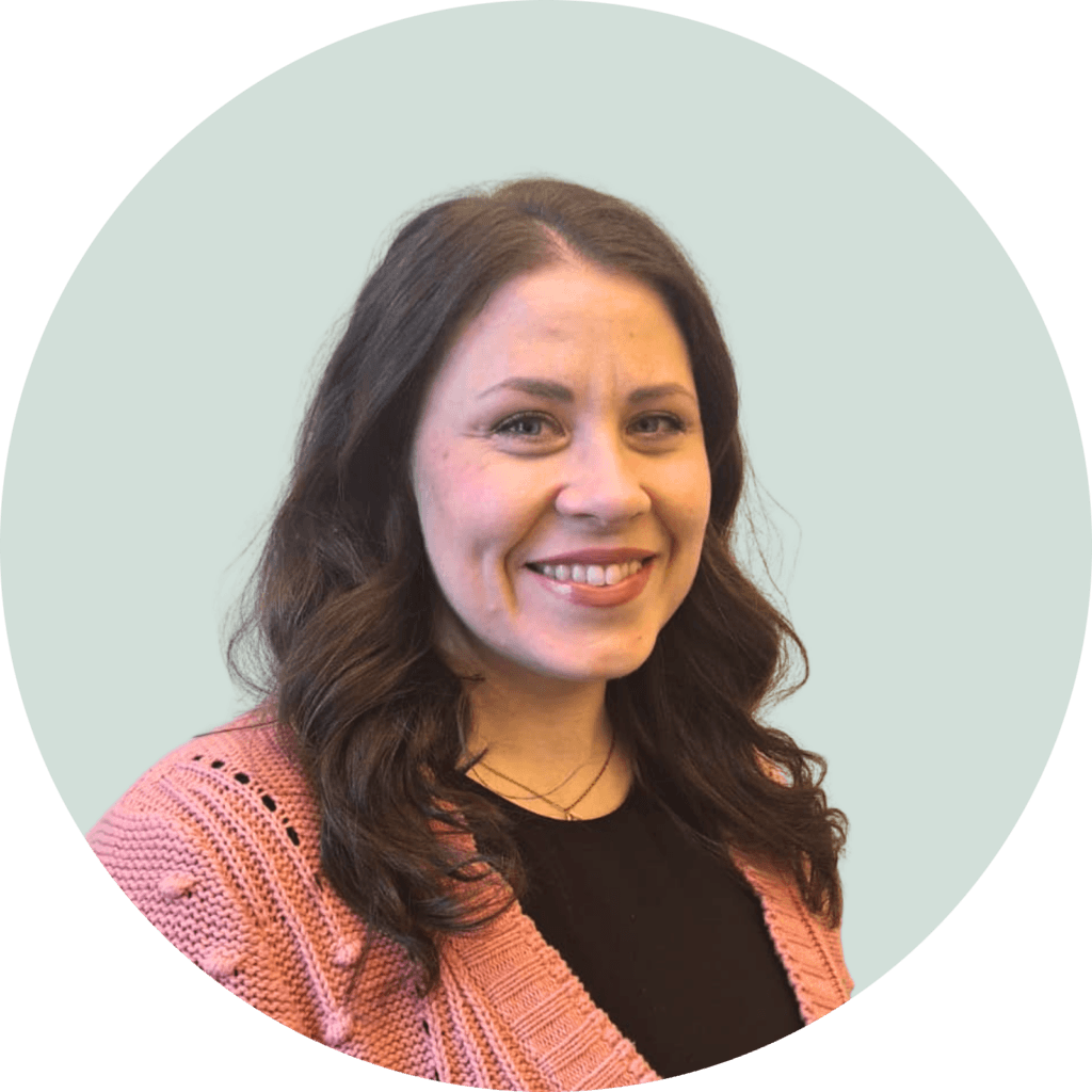 Hymyilevä henkilö vaaleansinisellä taustalla. Henkilöllä on ruskeat hiukset ja vaaleanpunainen neule.