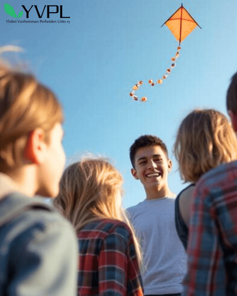 Nuori mies seisoo ulkona nauraen lasten kanssa, jotka katsovat häntä. Keskellä taivasta lentää oranssi leija, joka on koristeltu värikkäin koristein. Taustalla on kirkas sininen taivas. Kuvan vasemmassa yläkulmassa on YVPL-logo.