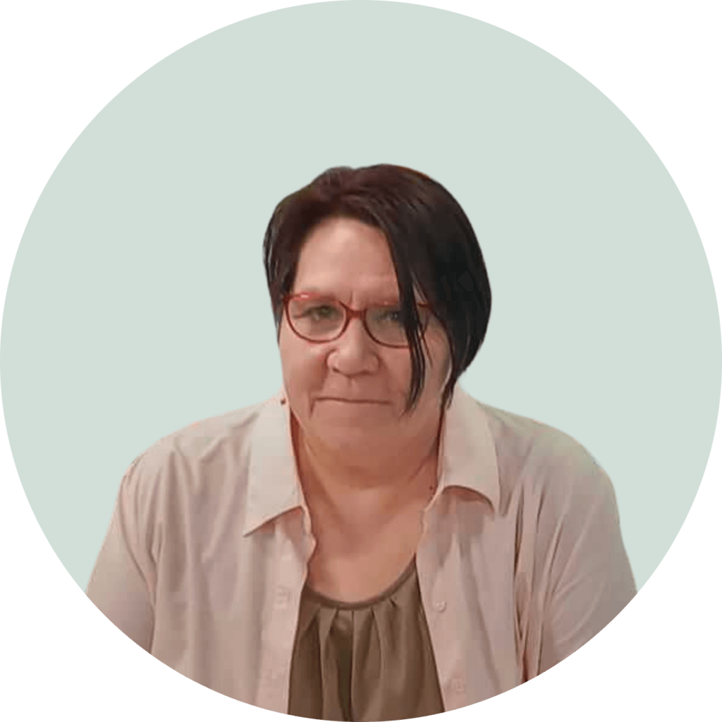 Alt text: Henkilö katsoo kameraan, hänellä on tummat hiukset ja punaiset silmälasit. Hänellä on päällään vaalea paita. Tausta on vaalean sinivihreä.