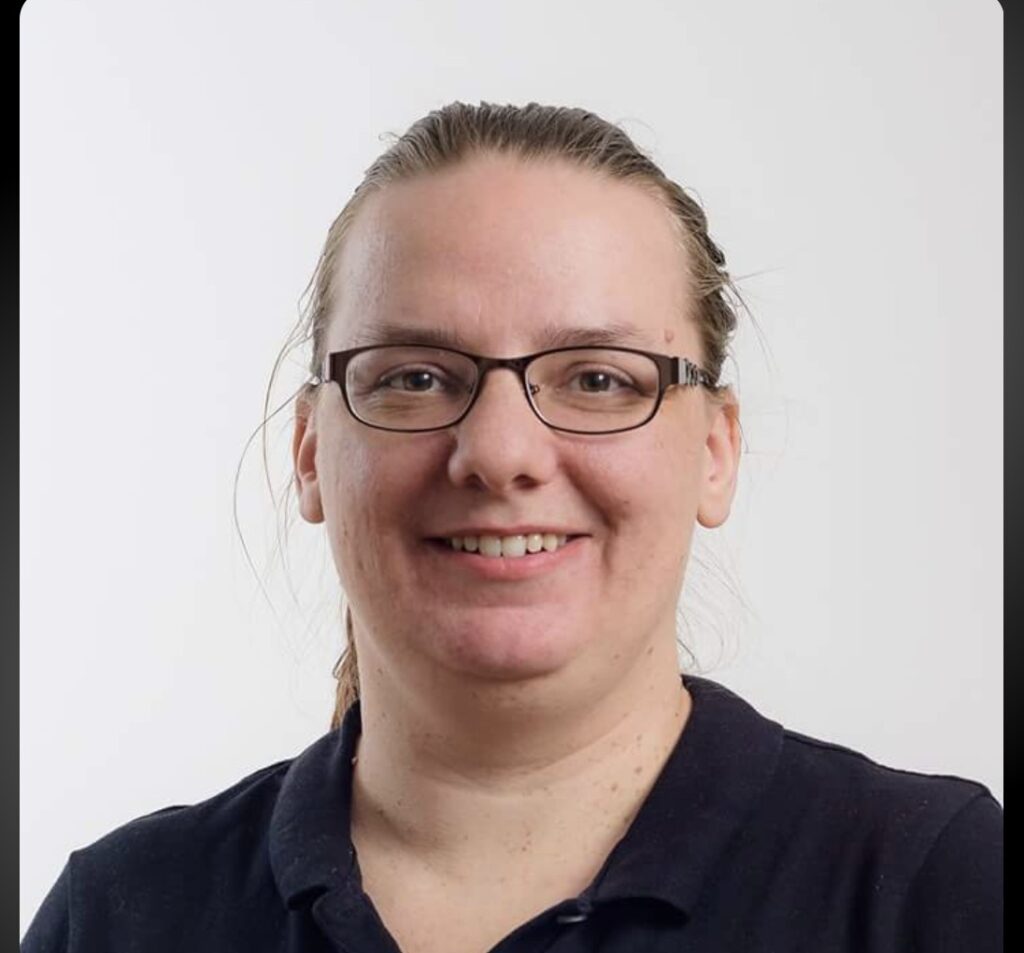 Kuvassa on henkilö, joka hymyilee. Hänellä on silmälasit ja yllään tummansininen paita. Tausta on vaalea.